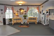 A cozy living room area with two wooden benches featuring floral cushions, a small coffee table with decorative items, and potted plants. The room has two windows with pink curtains, wall decorations including framed pictures and a cross, and a light fixture on the ceiling. The floor is carpeted with a small rug under the coffee table, and there is a partial view of a kitchen or service counter area on the right.