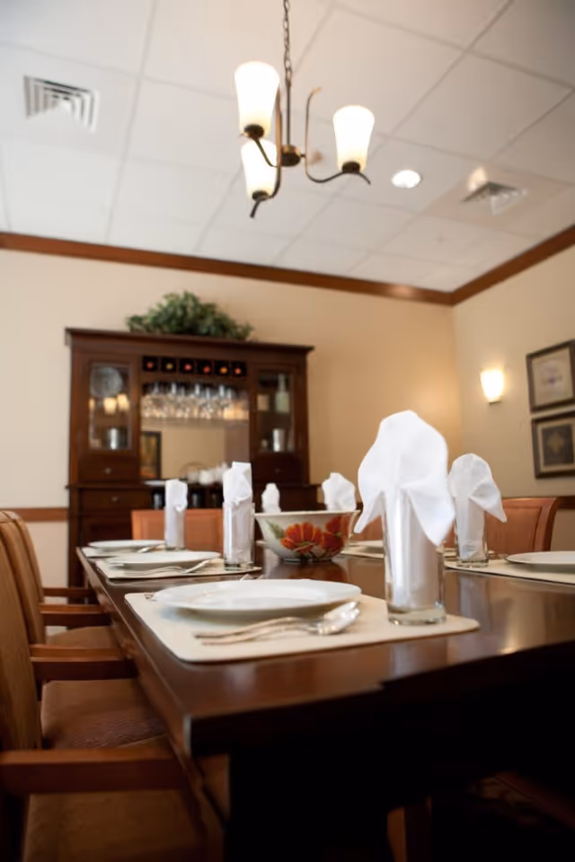 Formal dining room with a set wooden table, folded napkins in glasses, chairs, a decorative bowl, and a cabinet in the background.