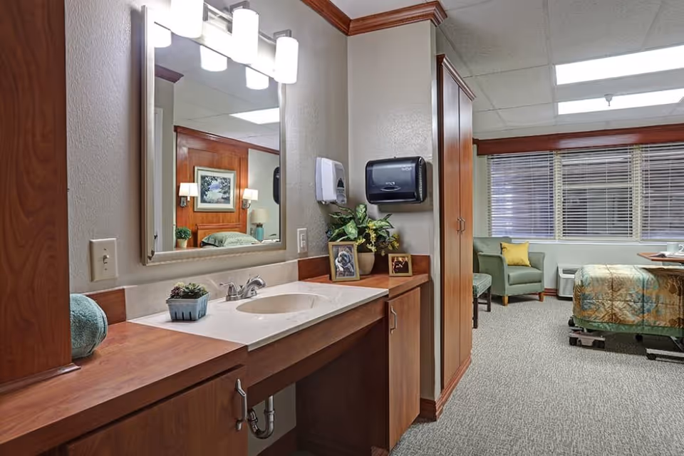 A senior living bedroom featuring a wood vanity with sink in the foreground and a bed and armchair by a window in the background.