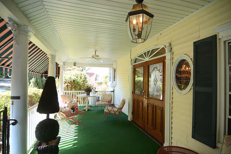 Covered porch area with green carpet, white columns, and a wooden double door with decorative glass panels. The porch has outdoor furniture including chairs and a small table with flowers, a ceiling fan, and a hanging lantern light fixture. There is a striped awning on the left side and some greenery visible outside.