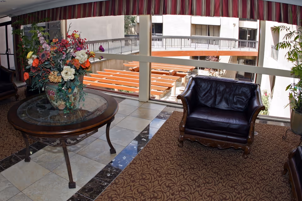A cozy indoor seating area with a dark leather armchair on a patterned carpet next to a round glass-top wooden table holding a large floral arrangement. Large windows behind the seating area reveal an outdoor courtyard with wooden benches and a walkway above.