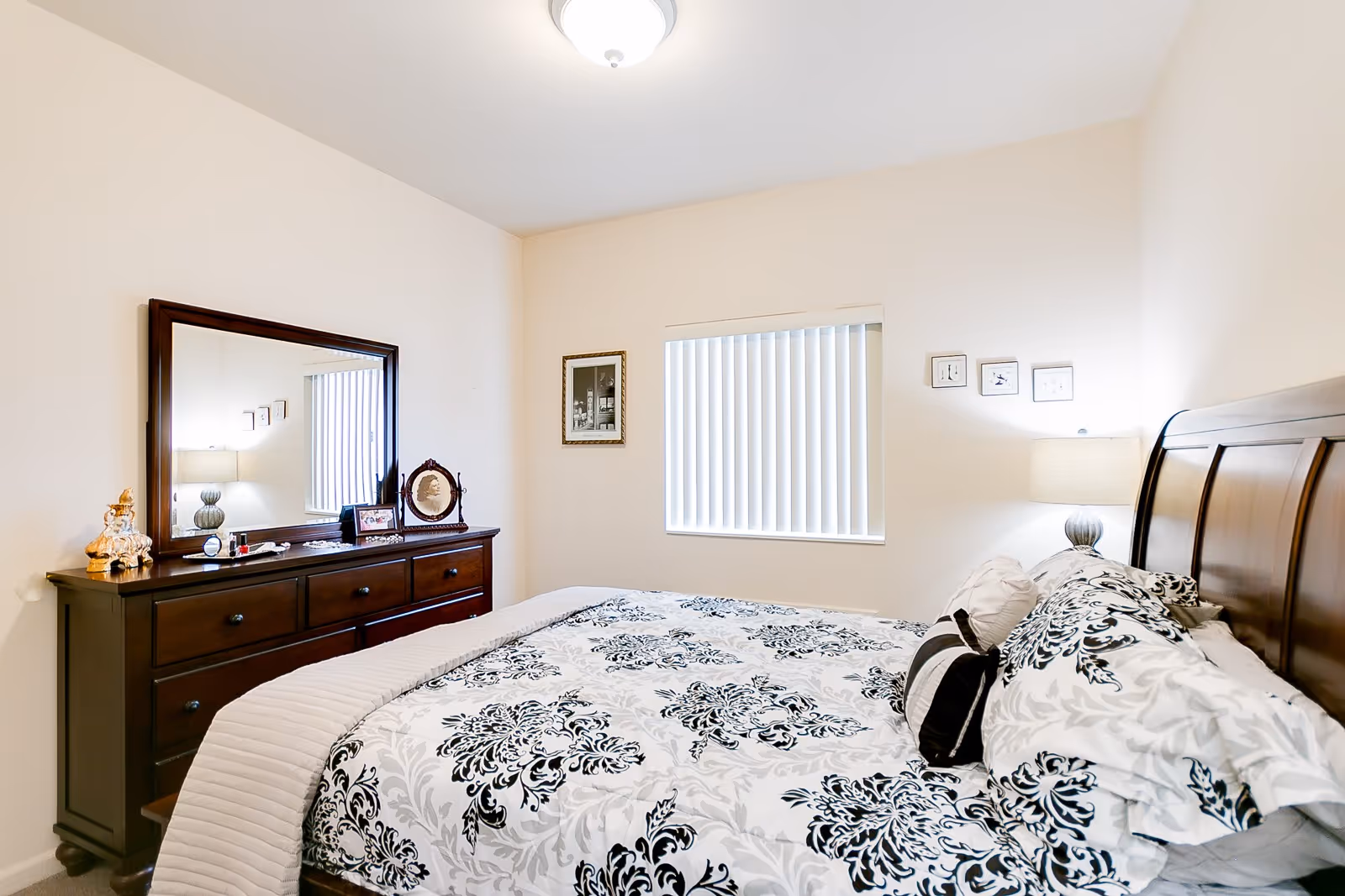 A bright bedroom with a large bed featuring a black and white floral patterned comforter and pillows. There is a wooden dresser with a large mirror on the left side, decorated with small framed pictures and ornaments. A window with vertical blinds is centered on the wall, and a table lamp is on a nightstand next to the bed. The walls are painted light beige.
