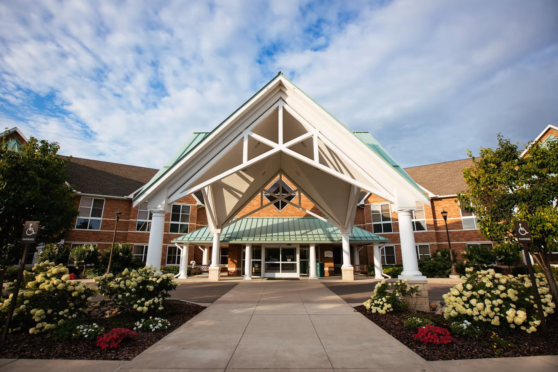 Front exterior view of Glacier Hills Senior Living Community building with a large covered entrance supported by white columns, landscaped flower beds with white and red flowers, and a partly cloudy sky overhead.