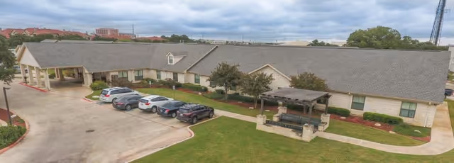 Exterior view of Sonterra Health Center showing a single-story building with a covered entrance, several parked cars, a small pergola with benches, and a well-maintained lawn under a cloudy sky.
