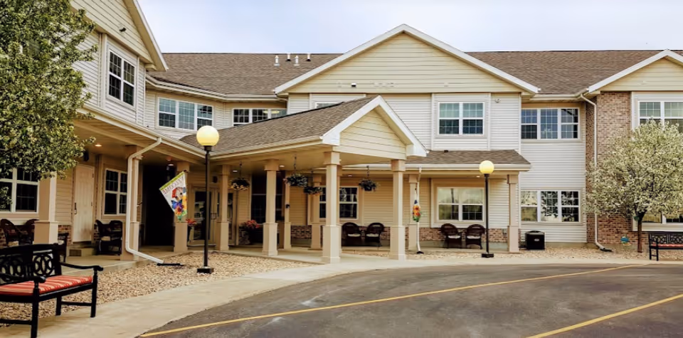 Exterior view of a senior living facility with beige siding and brick accents. The building has a covered entrance supported by columns, outdoor benches with red cushions, decorative hanging plants, and lamp posts. There are trees and a paved driveway in front.