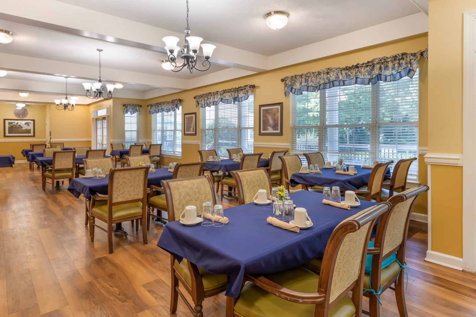 A dining room with multiple tables covered in blue tablecloths, each set with cups, glasses, and napkins. The room has wooden floors, yellow walls with white trim, large windows with floral valances, and several chandeliers hanging from the ceiling.