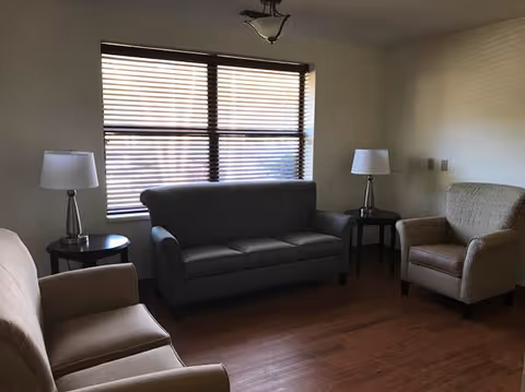 A small living room with a gray three-seat sofa centered under a window with wooden blinds. There are two beige armchairs, one on each side of the sofa. Each armchair has a round side table next to it with a silver lamp on top. The floor is wooden, and the walls are painted light beige.