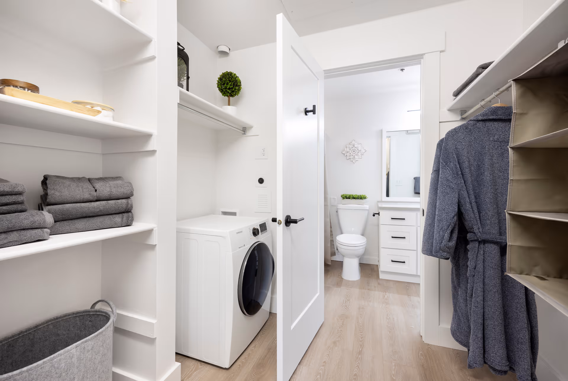 Bright walk-in laundry closet with a front-load washer, shelves of folded towels and a hanging robe, with an open door showing a bathroom toilet and vanity.