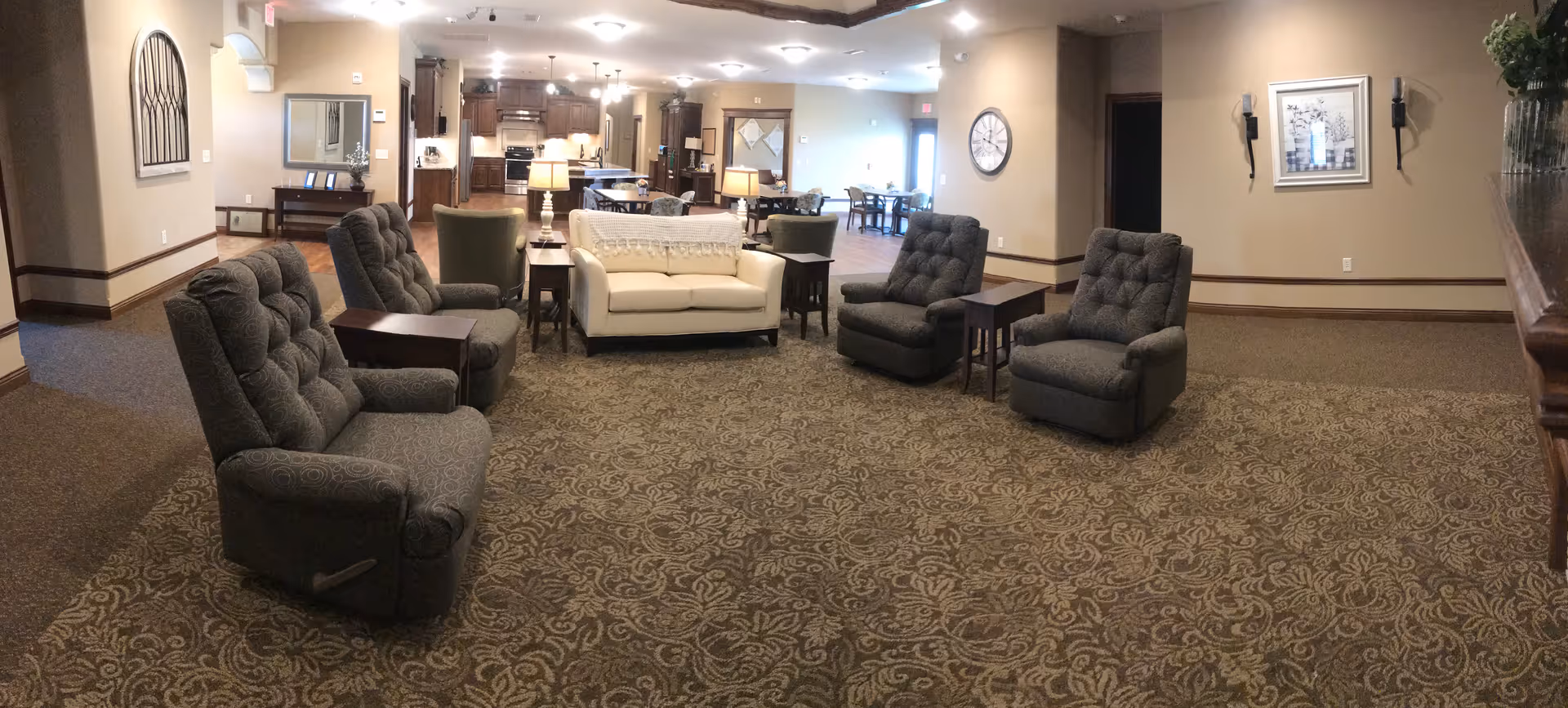 A spacious living area with patterned carpet, four gray recliner chairs, a white loveseat, and several wooden side tables with lamps. The background shows a kitchen and dining area with tables and chairs, beige walls, and decorative wall art and clocks.