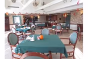 Dining area in a nursing home with multiple round and square tables covered with green tablecloths, surrounded by chairs. The room has a high ceiling with exposed beams and decorative hanging ornaments. There is a kitchen or serving area visible in the background.