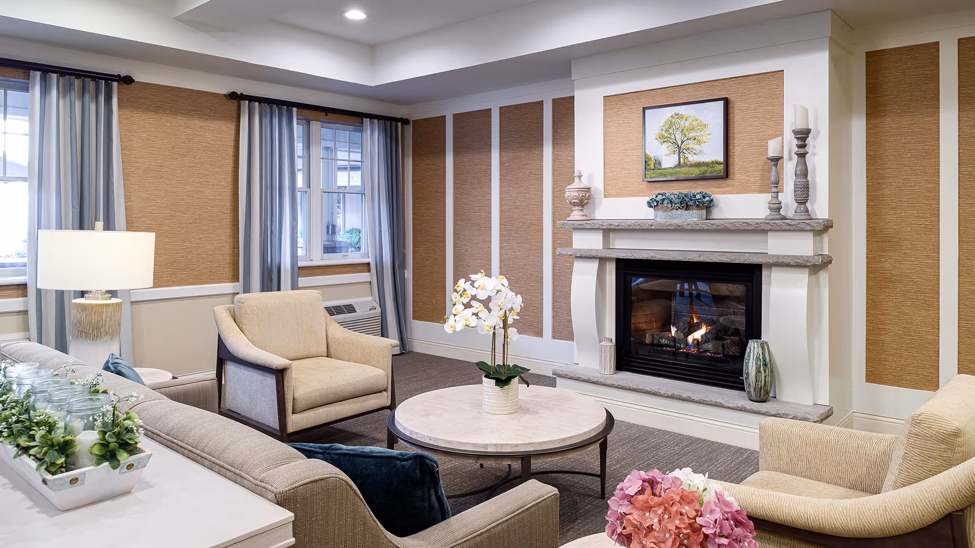 A cozy living room with beige armchairs and a sofa arranged around a round coffee table with a white orchid centerpiece. The room features a lit fireplace with decorative items and a framed tree painting above it. There are windows with gray curtains and a table lamp on a side table next to the sofa.
