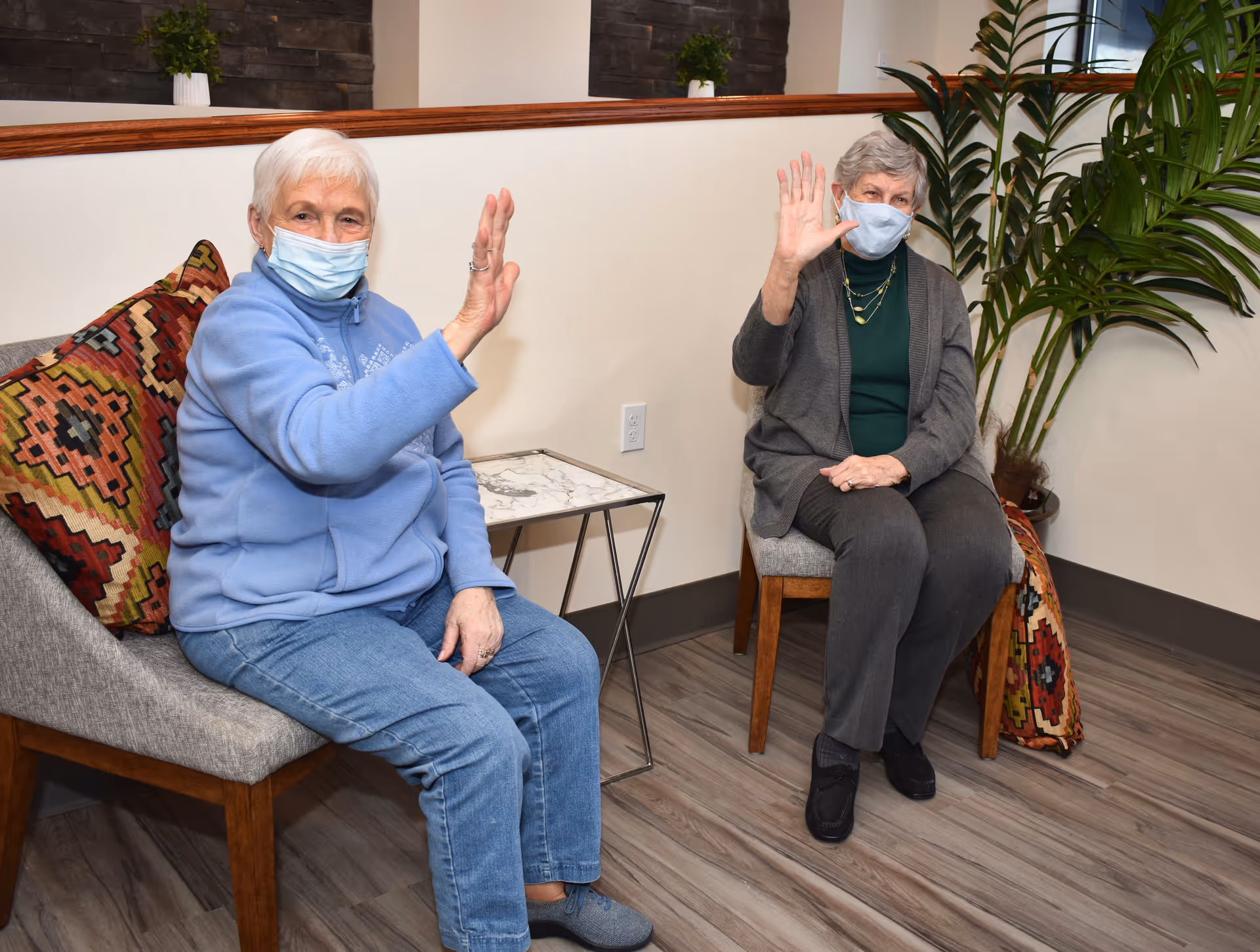 Two elderly women wearing face masks are sitting on chairs in a room with wooden flooring and a potted plant. They are both waving at the camera. One woman is wearing a blue jacket and jeans, while the other is dressed in a dark cardigan and pants. There is a small table with a marble top between them and decorative pillows on the chairs.