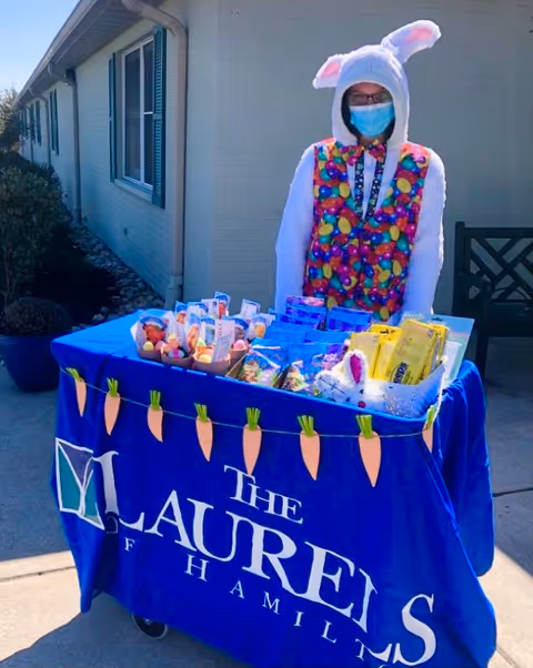 Person wearing a colorful vest and a white bunny hood with ears, along with a face mask, standing behind a table covered with a blue cloth that reads 'The Laurels of Hamilton'. The table is decorated with paper carrots and holds various packaged snacks and small baskets, set outdoors next to a building.