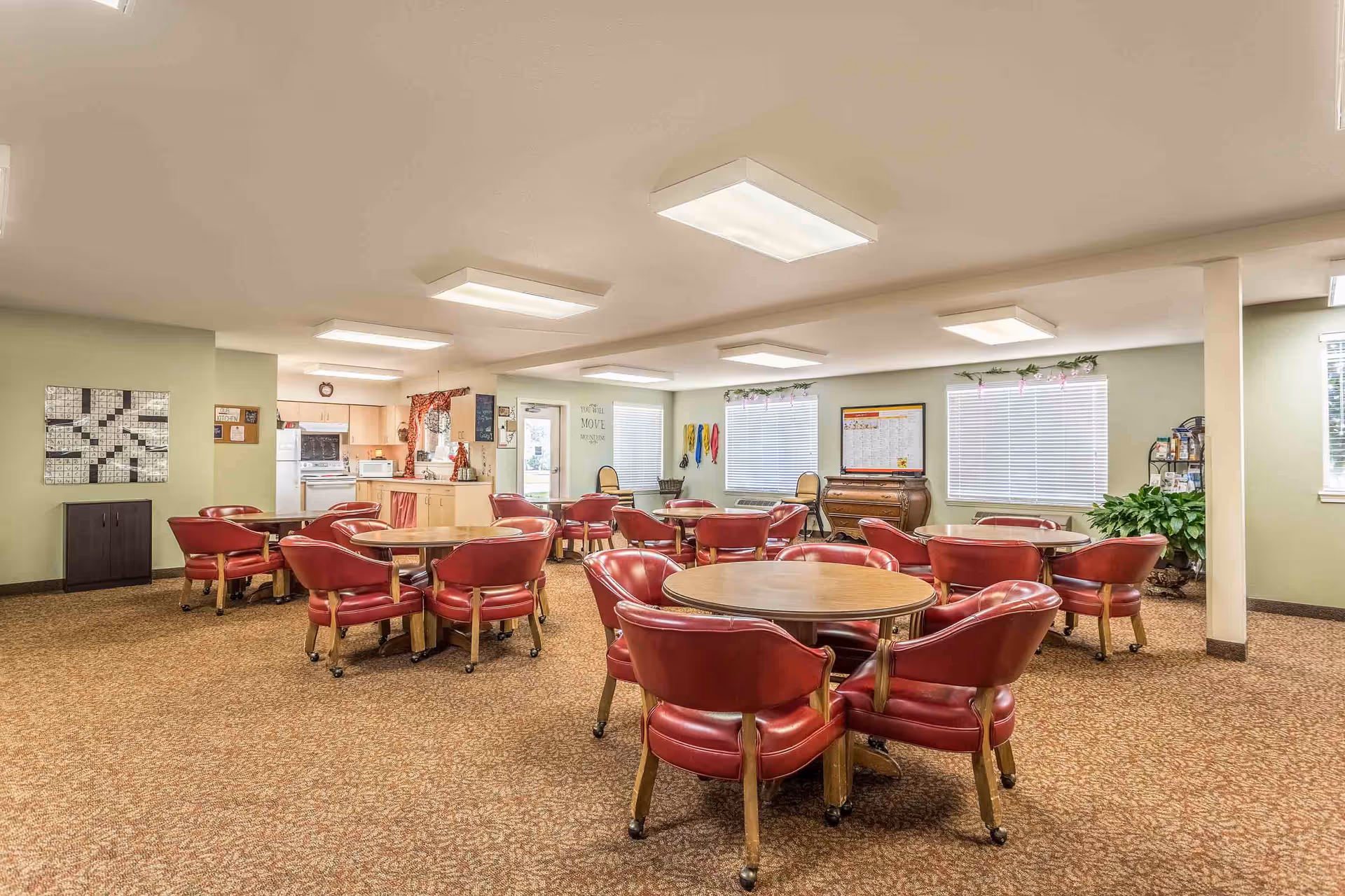 Communal dining room with round wooden tables and red upholstered chairs and a small kitchenette in the background.