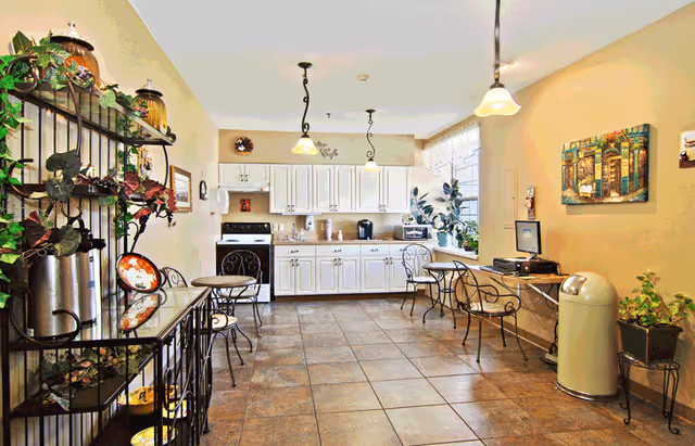 Bright communal dining and kitchenette area with white cabinets, small round tables and decorative plants.