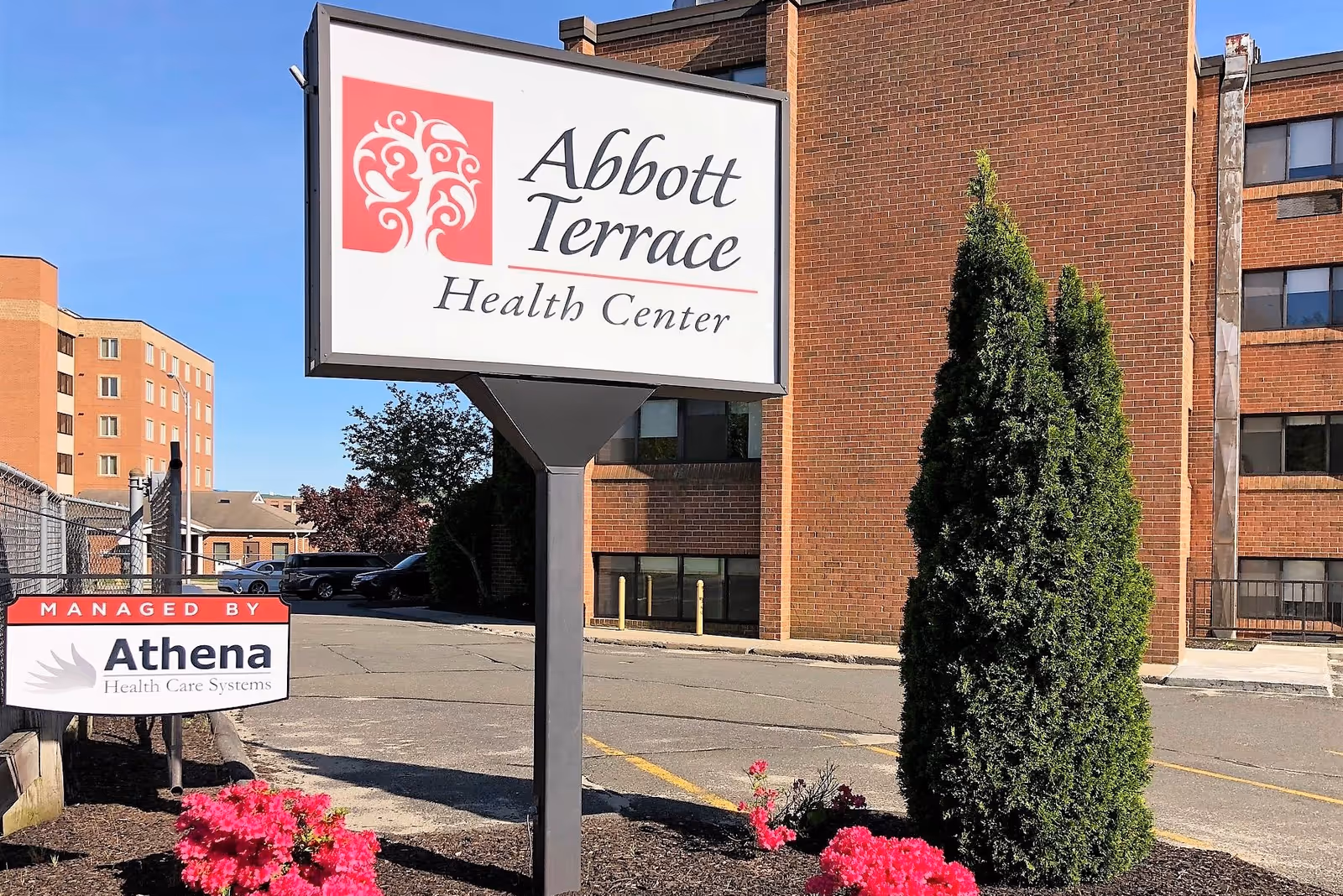 Outdoor view of Abbott Terrace Health Center sign mounted on a post with a brick building in the background, a small landscaped area with pink flowers and a tall green shrub, and a smaller sign indicating management by Athena Health Care Systems.