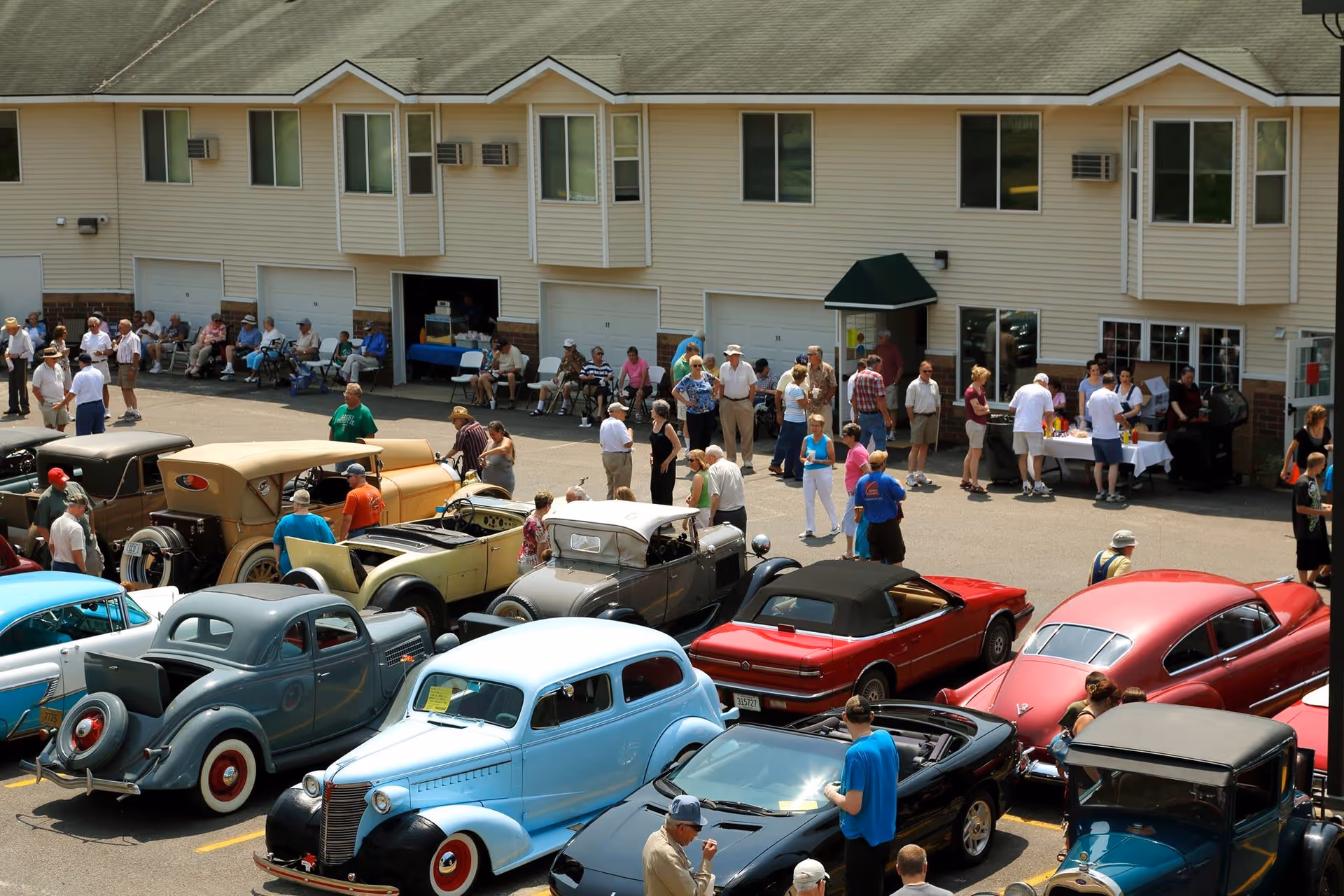 A group of people gathered outside a senior living facility named Meadow Lakes Senior Living, with several vintage and classic cars parked in the foreground. The building has multiple windows and garage doors, and people are socializing near a table with refreshments.