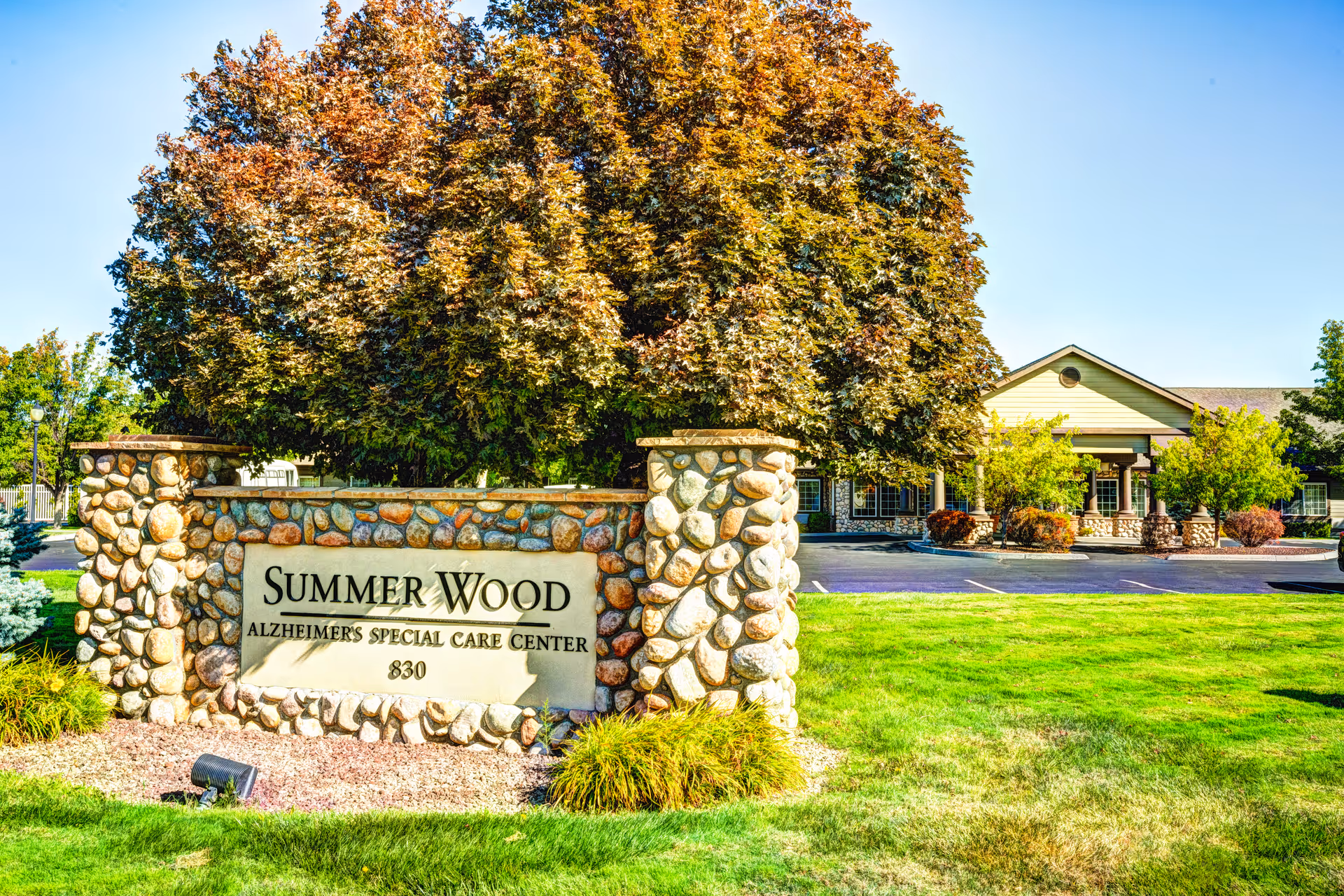 Stone sign for Summer Wood Alzheimer's Special Care Center with the number 830, set in a landscaped area with green grass and a large tree with autumn-colored leaves. In the background, there is a single-story building with a covered entrance and surrounding shrubs and trees under a clear blue sky.