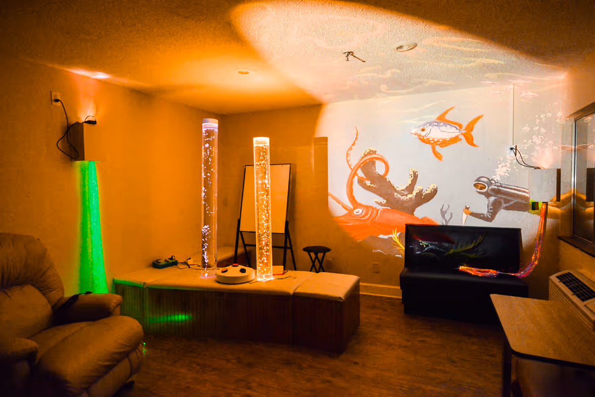A dimly lit sensory room with underwater-themed projections on the wall, including a diver, fish, and an octopus. The room features two tall bubble tubes emitting light, a comfortable recliner chair, a black cushioned bench, and a wooden table. The floor is wooden, and there are some small stools and a whiteboard in the background.
