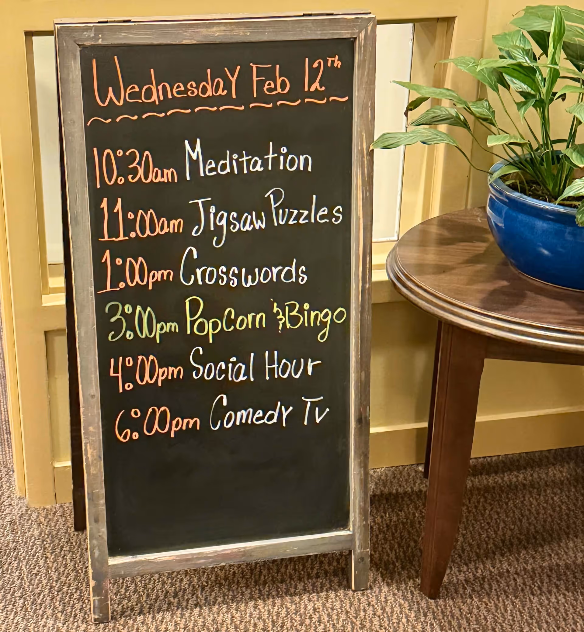 Chalkboard sign listing daily activities and times next to a round table with a potted plant.