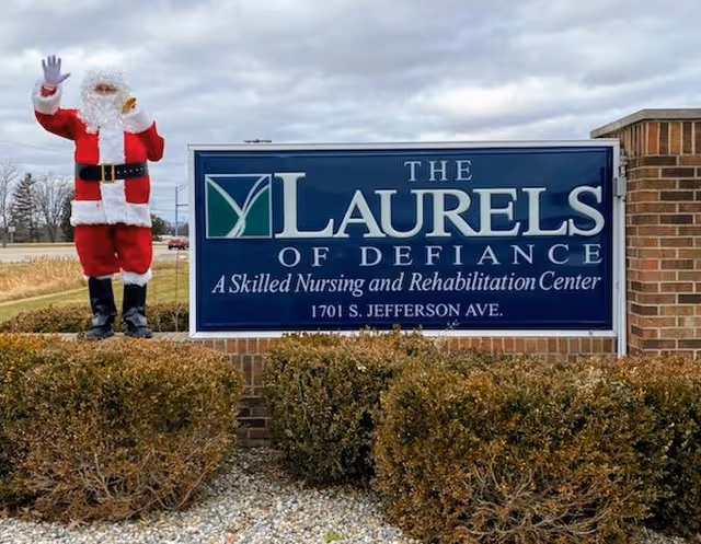 A large blue sign for The Laurels of Defiance, a skilled nursing and rehabilitation center located at 1701 S. Jefferson Ave. Next to the sign stands a person dressed in a Santa Claus costume waving. The sign is mounted on a brick base with bushes and gravel in front, and a cloudy sky in the background.