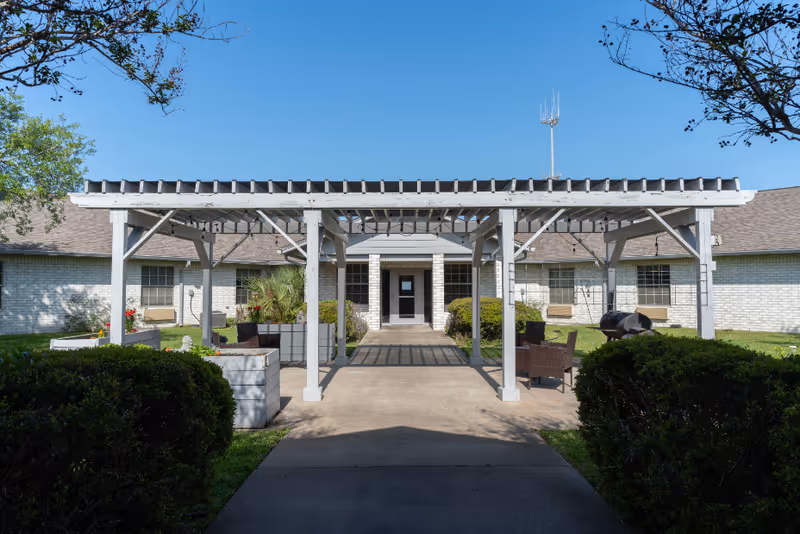 Outdoor view of a white pergola structure with seating and planters in front of a single-story brick building under a clear blue sky. The building has multiple windows and a central entrance door.