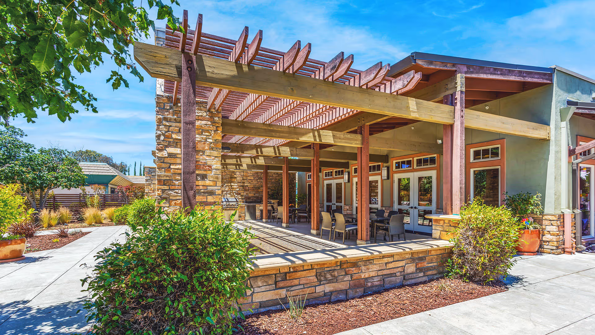 Outdoor patio area at Atria Valley View featuring a wooden pergola with beams casting shadows on the ground, stone pillars, and a seating area with tables and chairs. Surrounding the patio are landscaped plants and trees under a clear blue sky.