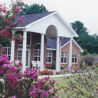 Front exterior view of a brick building with white columns and a covered entrance. There are white chairs on the porch and flowering bushes with purple and pink blooms in the foreground. Trees and a wooden fence are visible in the background.
