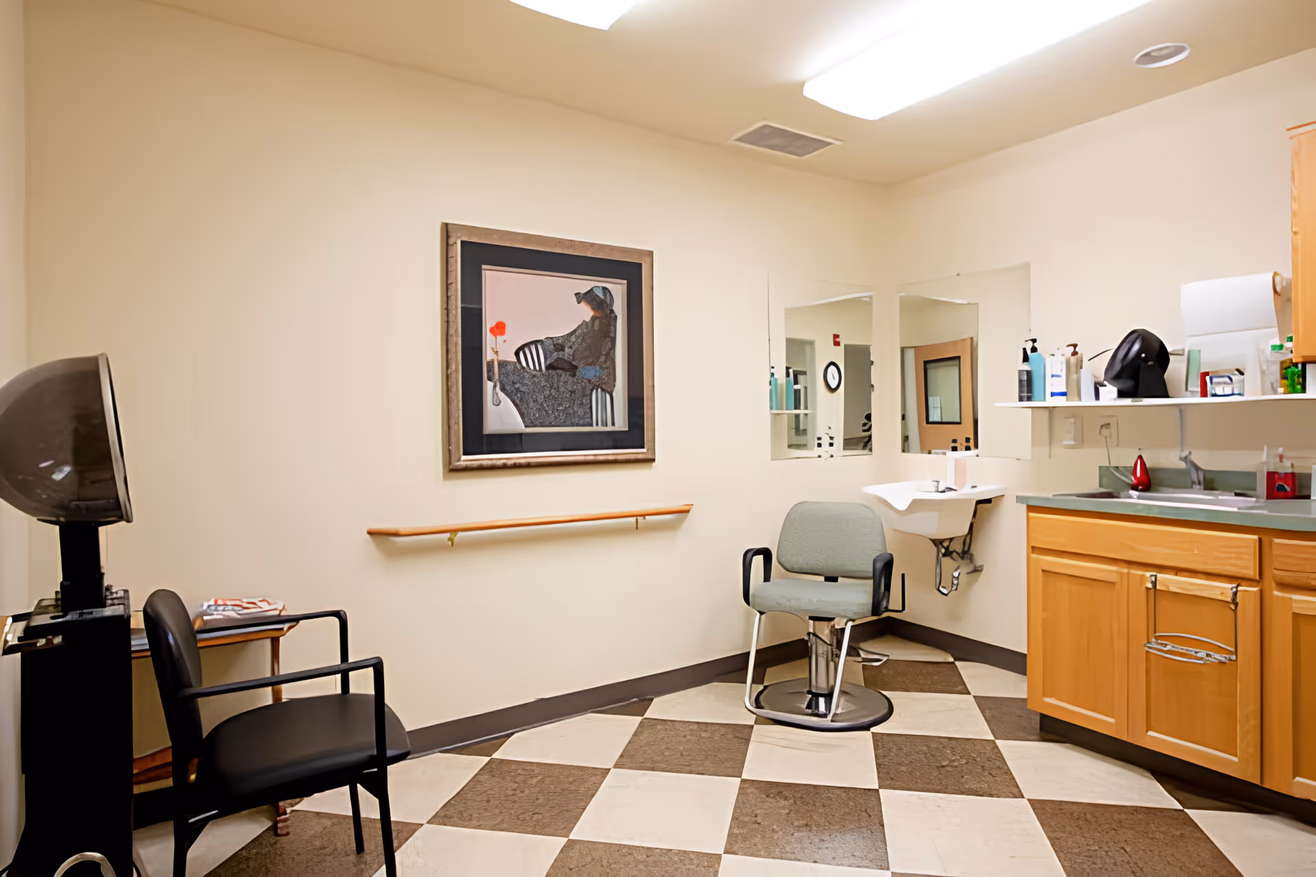 Interior of a senior living facility hair salon with a salon chair, a black waiting chair, a hair dryer, a sink, cabinets, and a framed artwork on the wall. The floor has a checkered pattern with brown and beige tiles.