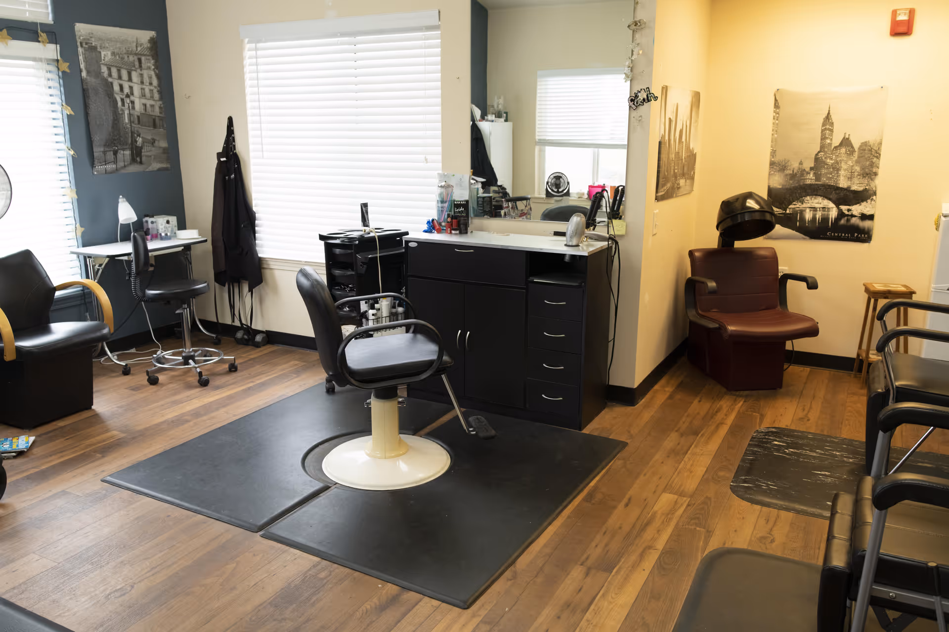 Interior of a hair salon area with a central black salon chair on a black mat, a black cabinet with a large mirror above it, and various salon tools on the counter. The room has wooden flooring, a window with white blinds, and several black chairs around the perimeter. There are black and white cityscape posters on the walls and a hair dryer chair in the corner.