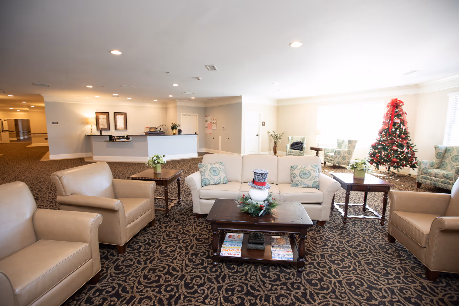 Spacious assisted-living lobby with sofas and armchairs arranged around a coffee table, a reception desk in the background, and a decorated Christmas tree to the right.