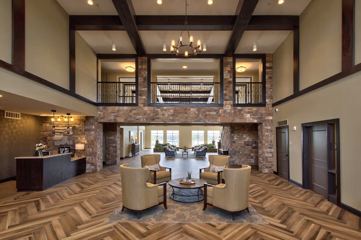 Spacious senior living facility common area with high ceilings, exposed wooden beams, and a chandelier. The room features a seating area with four beige armchairs around a round coffee table on a patterned rug. There is a reception desk to the left with flowers and framed photos, and a stone accent wall behind it. Large windows in the background let in natural light, illuminating additional seating areas with sofas and plants.