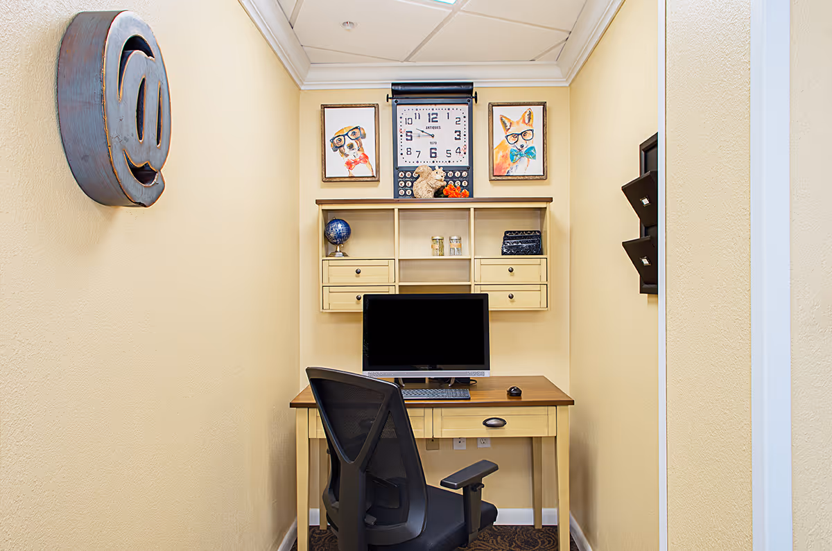 Small office nook with a desk, computer monitor and swivel chair beneath wall shelves and framed artwork.