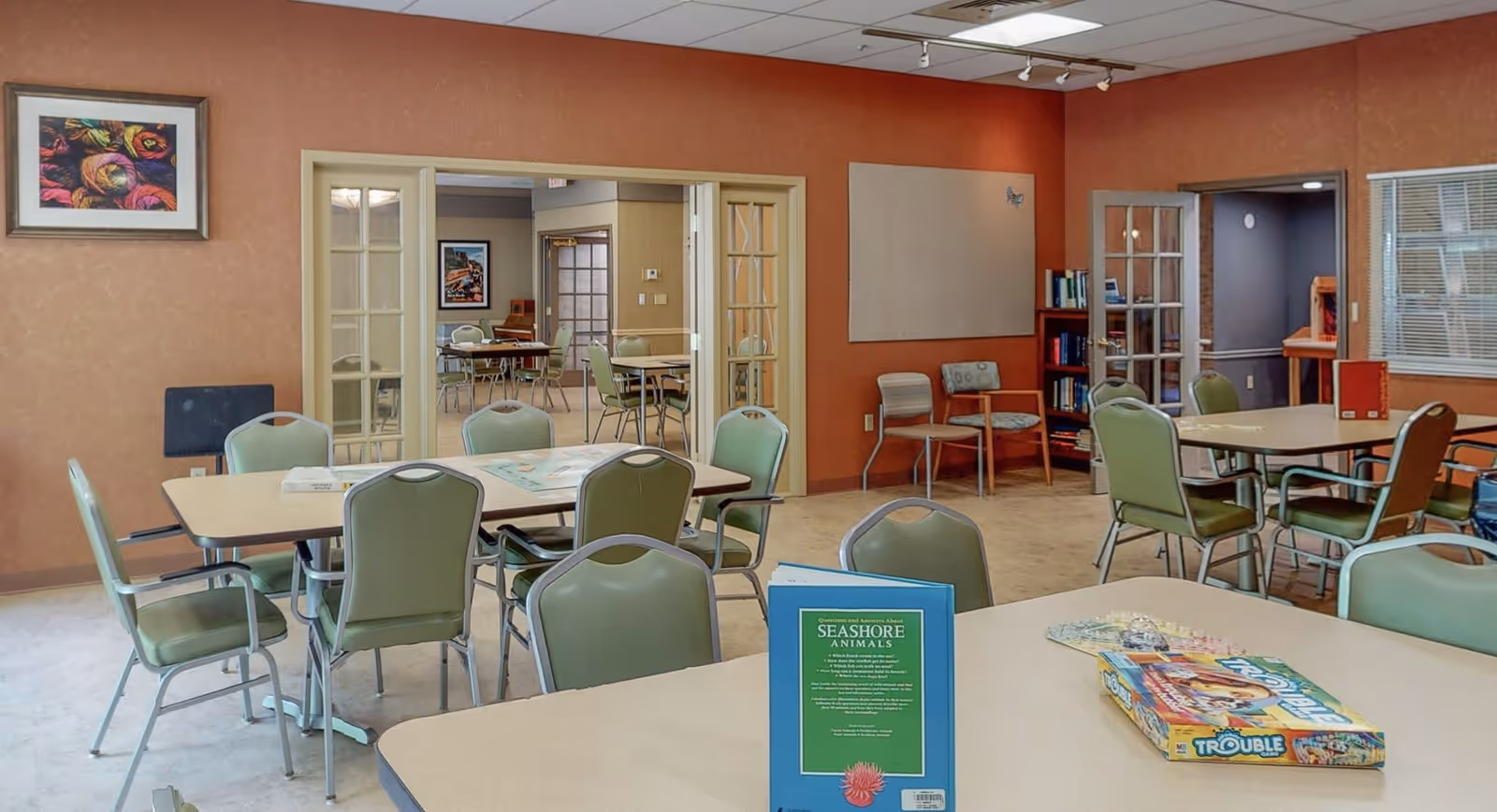 A communal activity room with several tables and green chairs arranged around them. On the closest table, there is a book titled 'Seashore Animals' and a board game called 'Trouble'. The room has warm-toned walls, framed artwork, and a bookshelf with books. There are glass-paneled double doors leading to another room with more tables and chairs.