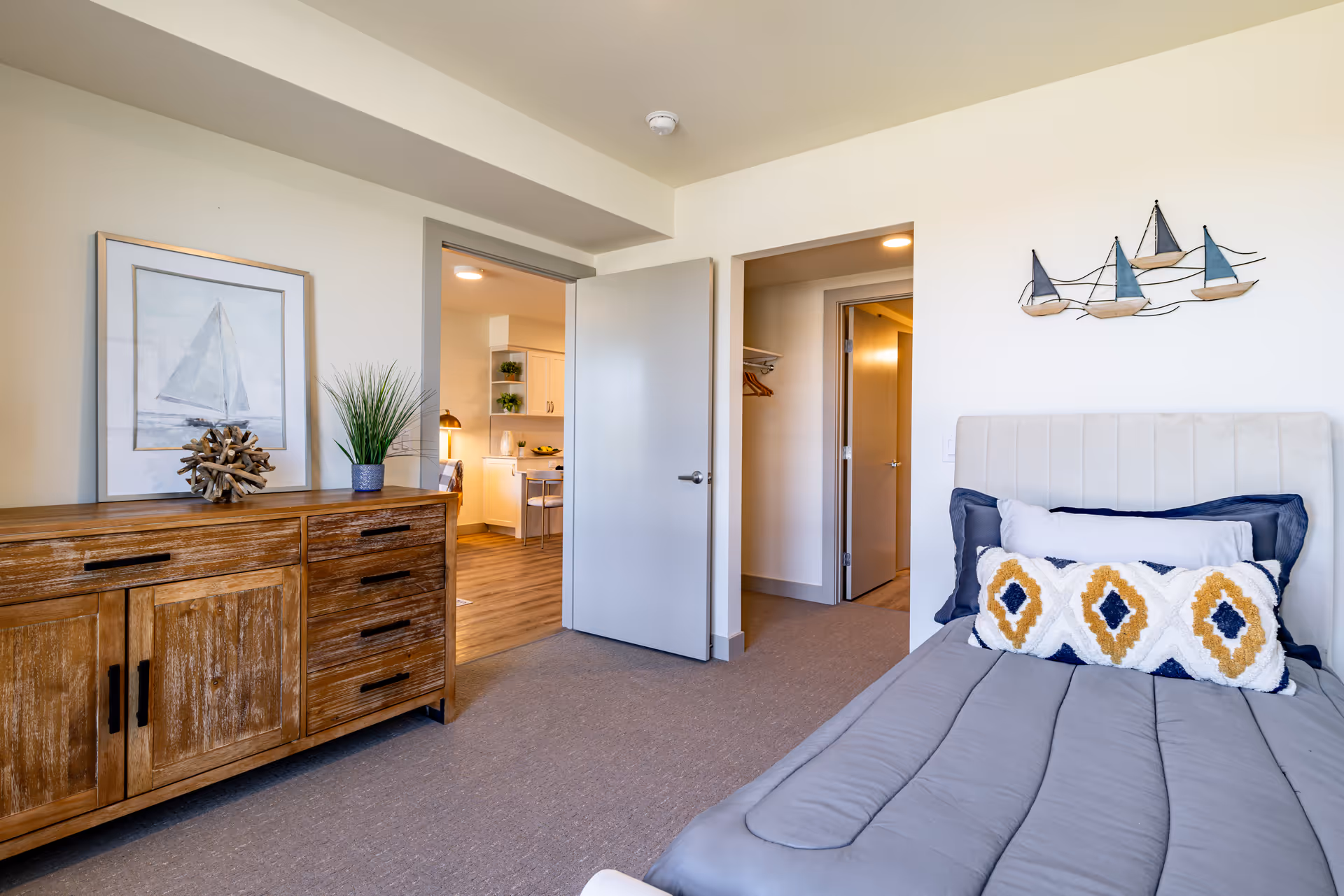 Furnished bedroom with a bed and decorative pillows, a wooden dresser, and an open door revealing a kitchen and hallway.