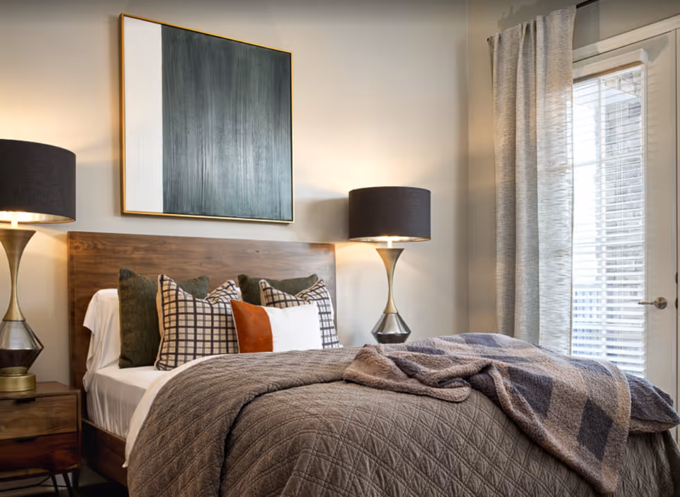 A cozy bedroom with a wooden bed frame, multiple pillows in various patterns and colors, a textured gray blanket, two bedside tables each with a modern lamp, a large abstract painting above the bed, and a window with blinds and curtains allowing natural light to enter.