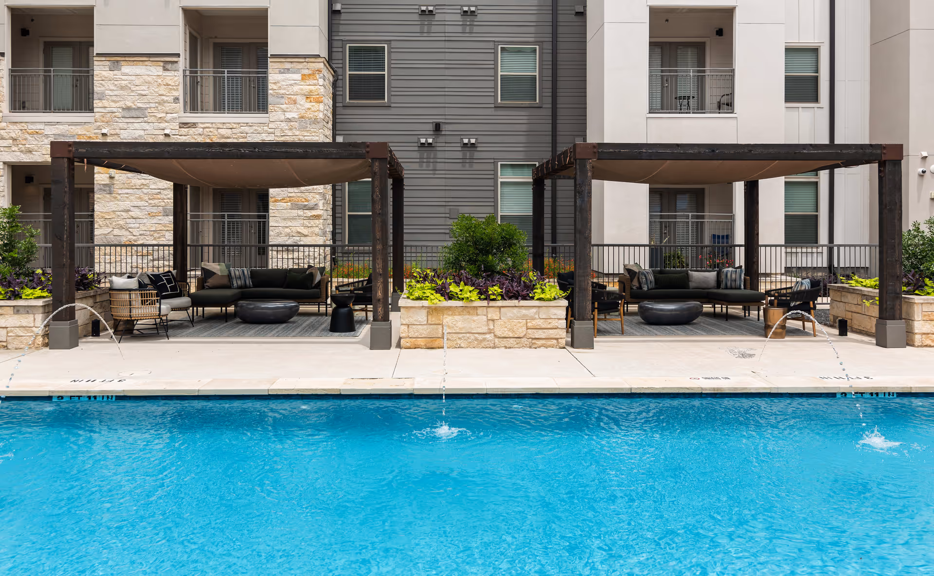 Outdoor pool area with two shaded seating cabanas featuring cushioned chairs and sofas, surrounded by stone planters with green and purple plants, in front of a multi-story building with balconies and windows.