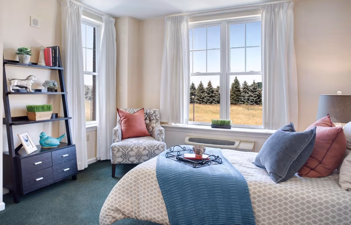 A cozy bedroom with a large window showing a view of trees and a field outside. The room features a bed with multiple pillows and a blue throw blanket, a patterned armchair with a red pillow, and a black shelving unit holding decorative items including a small plant, books, and a turquoise bird figurine. White curtains frame the windows, and the carpet is green.
