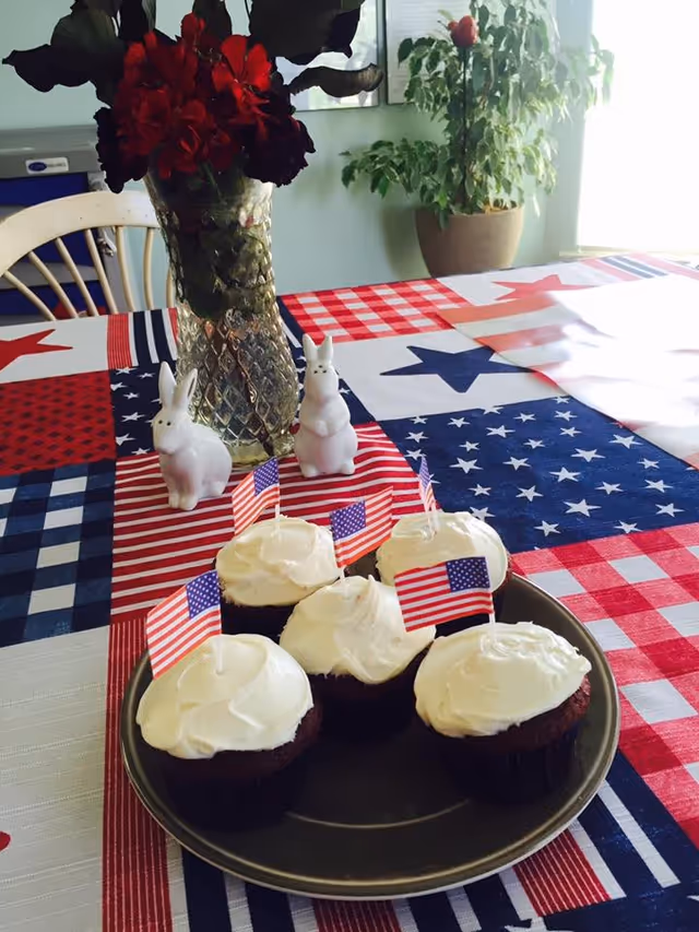 A table decorated with a patriotic red, white, and blue tablecloth featuring stars and stripes. On the table is a plate with five cupcakes topped with white frosting and small American flags. Behind the cupcakes, there is a glass vase with red flowers and two white ceramic bunny figurines. In the background, a potted plant is visible near a window.
