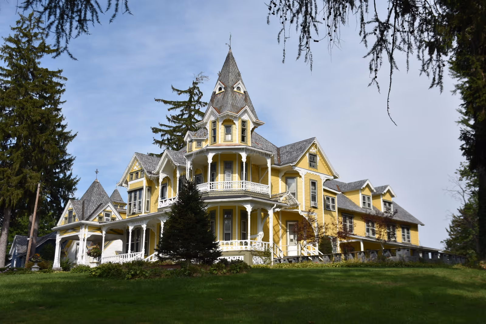 Large yellow Victorian-style multi-story house with a wraparound porch and tower on a grassy lawn.