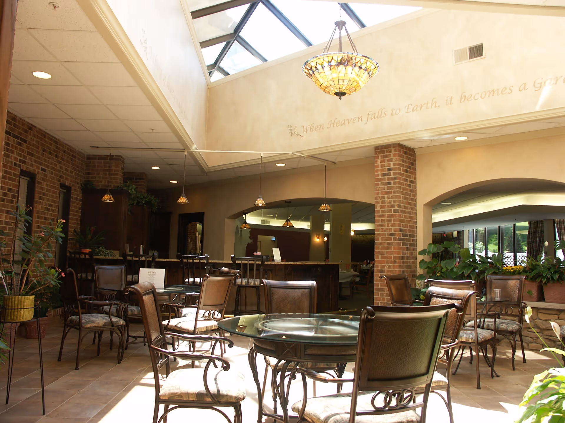 A bright indoor seating area with glass-top tables and cushioned chairs arranged on a tiled floor. The space features brick columns, hanging pendant lights, and a large skylight ceiling allowing natural light to flood in. There are plants around the perimeter and a partial view of another room through an archway.