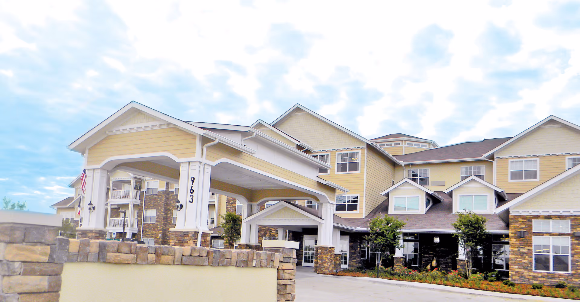 Exterior view of Liberty Heights Gracious Retirement Living facility showing a large building with yellow siding, stone accents, multiple windows, and a covered entrance with the number 963 displayed on a pillar. The sky is partly cloudy.