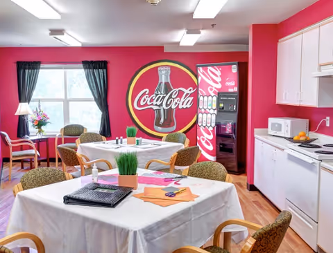 Bright dining/activity room with round tables and chairs, a large Coca-Cola mural and vending machine, and a small kitchenette.