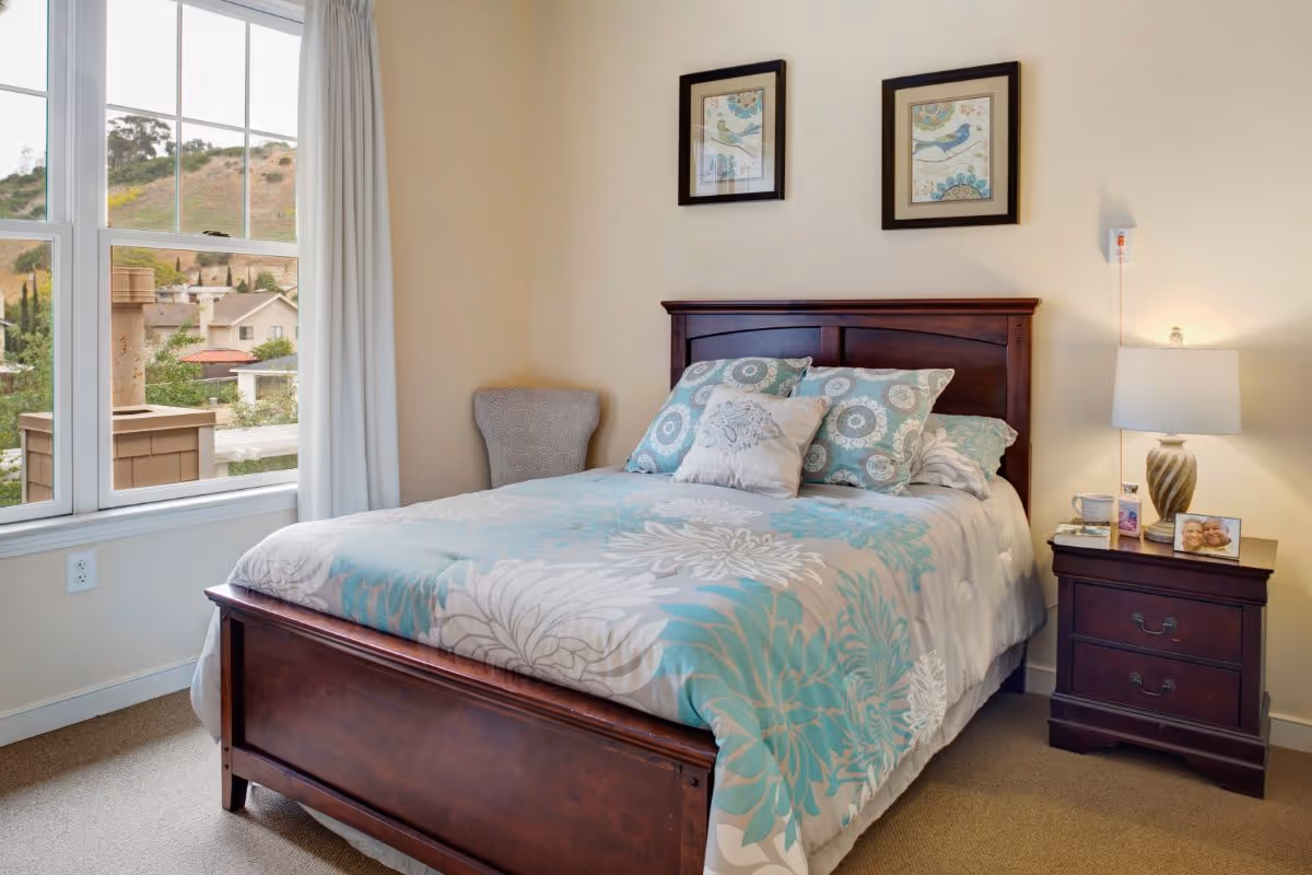 A cozy bedroom with a wooden bed frame and a bed made with a floral patterned comforter in shades of blue, white, and gray. There are multiple pillows on the bed. Next to the bed is a wooden nightstand with a lamp, a framed photo, and some small items. Two framed pictures hang on the wall above the bed. A window with white curtains shows a view of houses and hills outside.