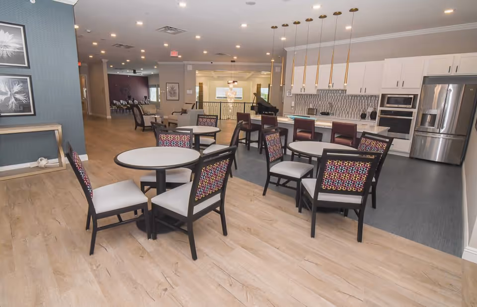 Open communal dining area with round tables and colorful upholstered chairs in front of a modern kitchen island with pendant lights.