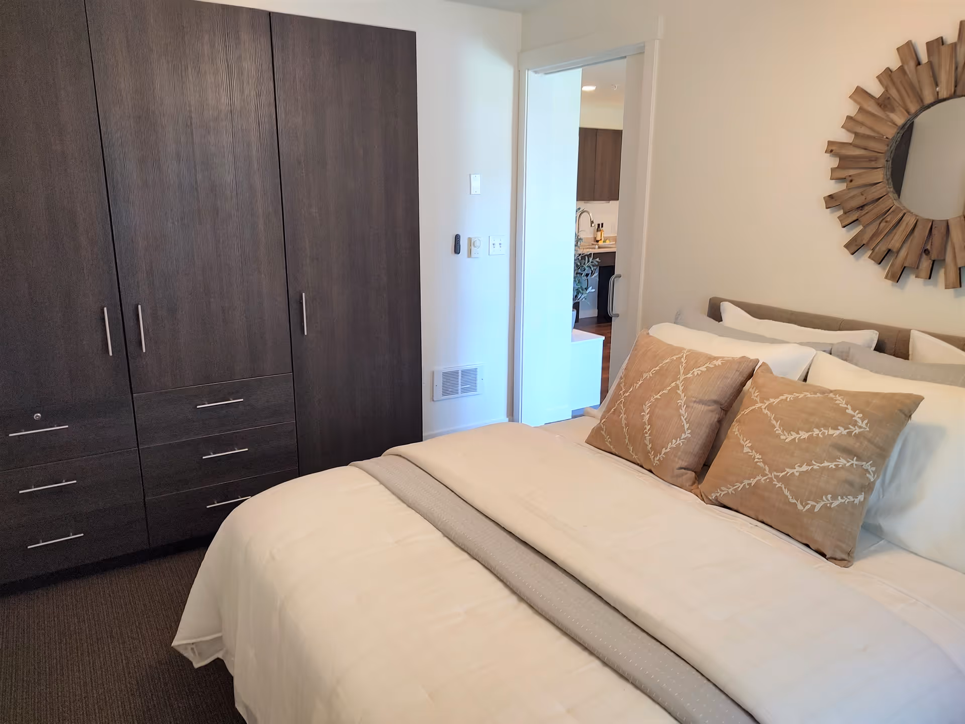 Well-lit bedroom with a made bed and decorative pillows, a large dark wardrobe, and a round wooden mirror on the wall.