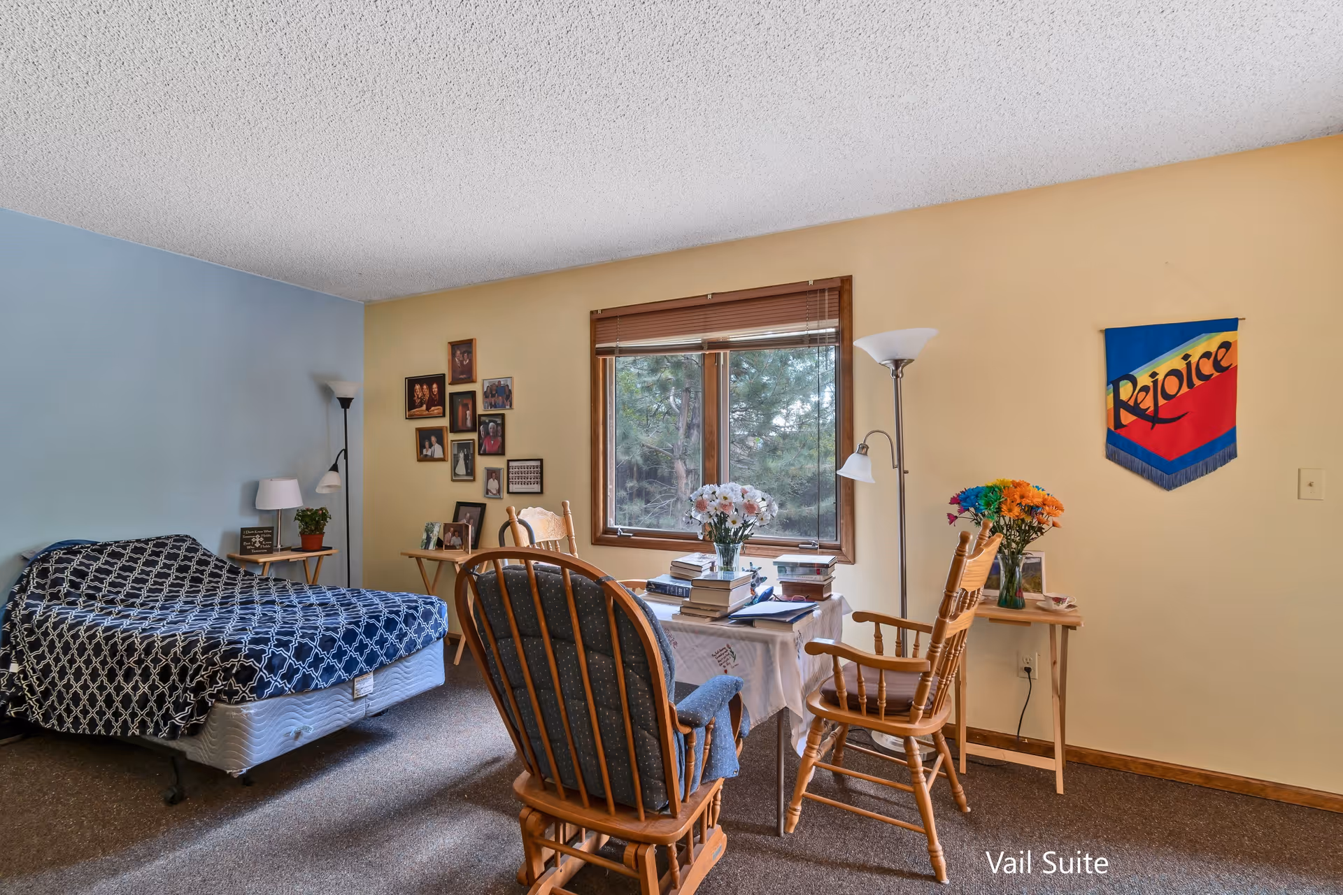 A cozy room with a single bed covered in a blue and white patterned blanket, a small table with a lamp and plant, a window with a view of trees, two wooden chairs around a small table with books and flowers, framed photos on the wall, and a colorful wall hanging that says 'Rejoice'.