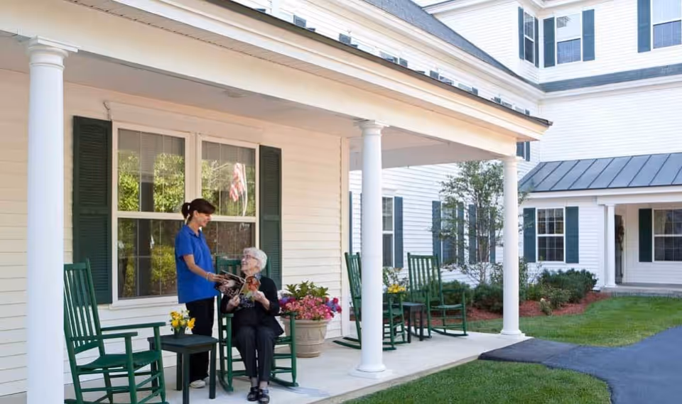 An elderly woman sits on a porch rocker while a caregiver stands beside her showing a magazine in front of a white senior living building.