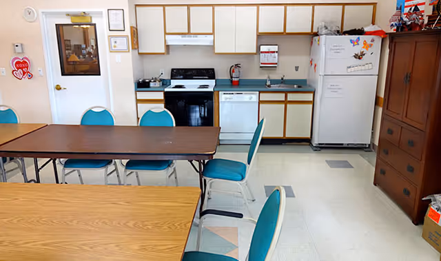 A kitchen and dining area in an assisted living facility with a stove, dishwasher, refrigerator, cabinets, and several tables with teal chairs arranged around them. There is a wooden cabinet on the right side and a door with a window on the left side of the room.