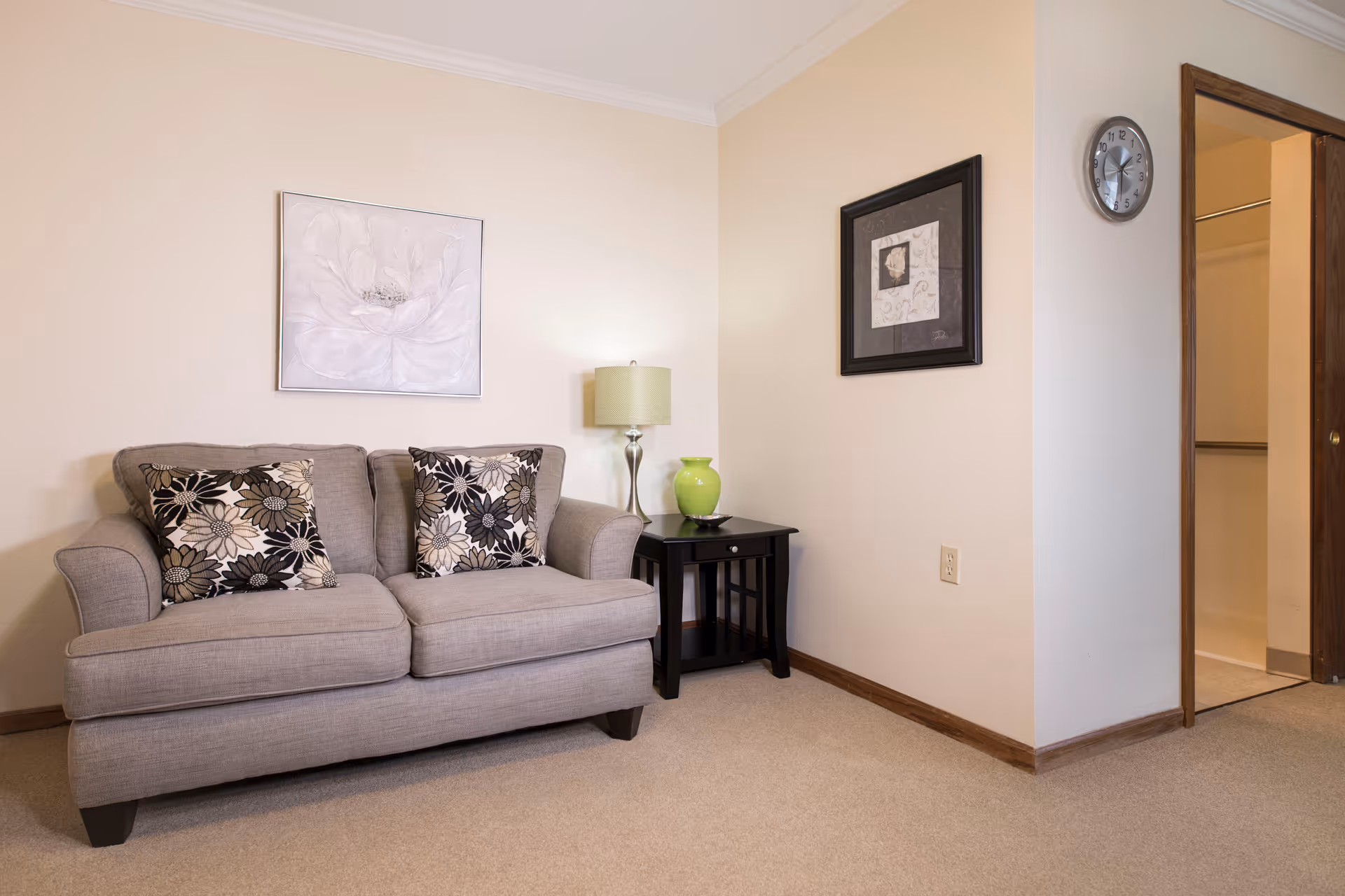 A small living room with a gray loveseat topped with patterned pillows, a side table holding a lamp and green vase, wall art, and an open doorway.